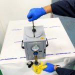 bulk hot melt handgun held underneath spigot of the gray premelt tank with the person's other hand on top the piston at the top of the premelt tank